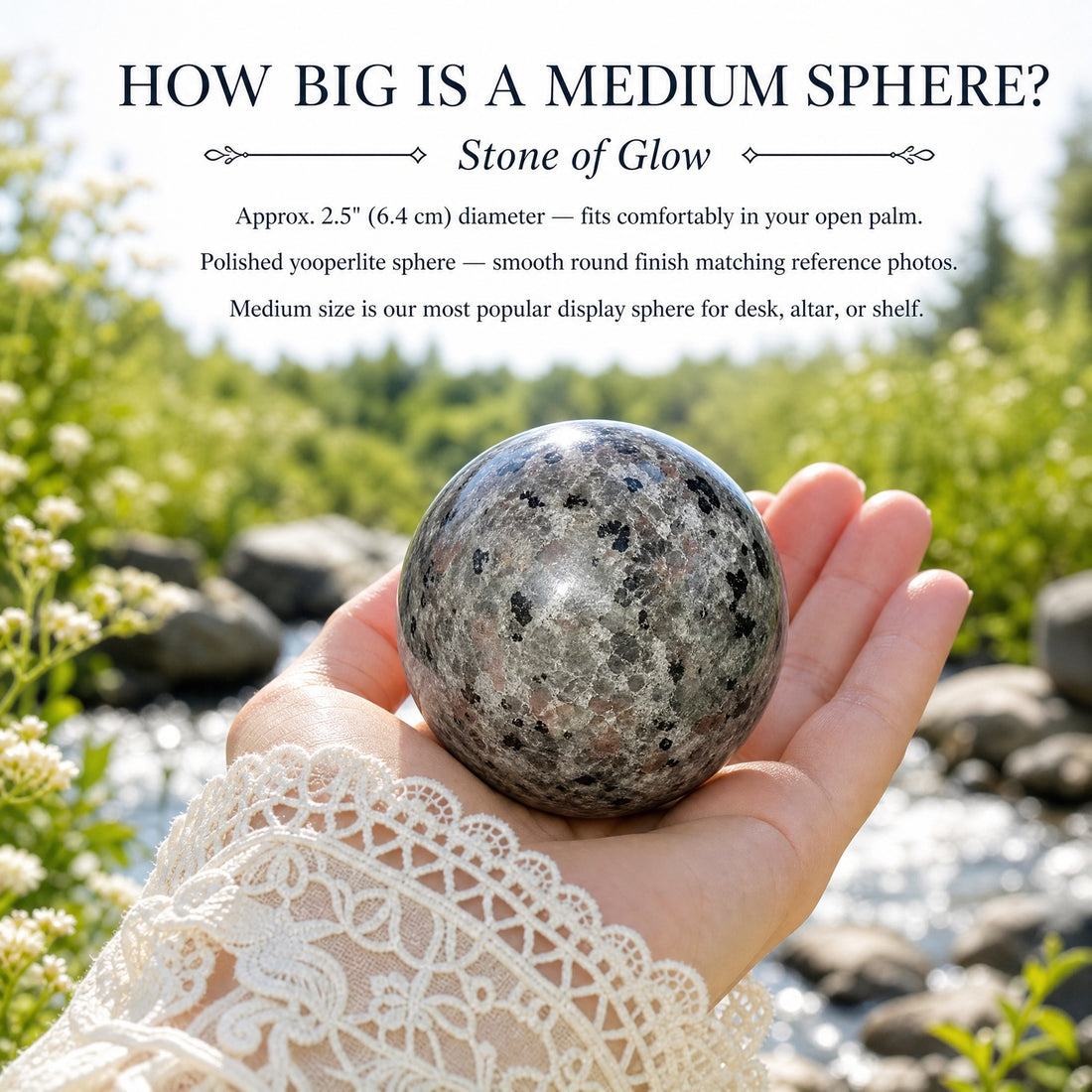 A polished gray yooperlite sphere speckled with black, resting in an open palm beside a stream