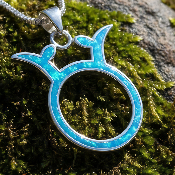 Close-up of the Taurus glyph pendant, a circle crowned with curved horns, inlaid with blue opal in sterling silver, resting on moss