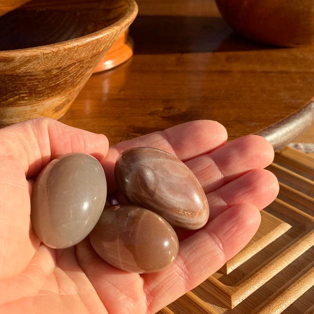 Three polished golden sunstone ellipses in warm peach and taupe tones resting in an open palm in sunlight