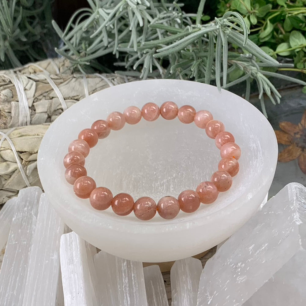 A peach and golden sunstone bead bracelet resting in a white selenite bowl beside sage bundles