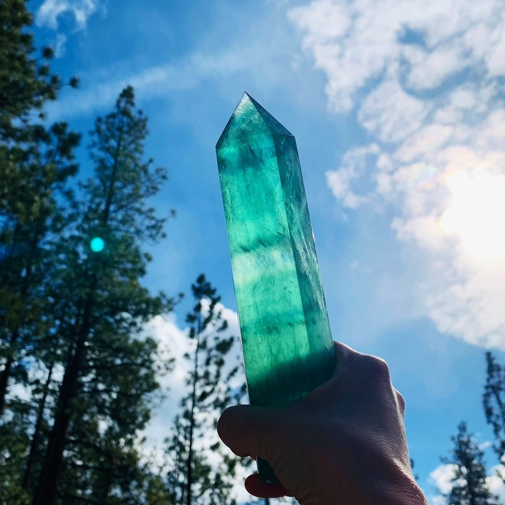 A large green fluorite generator held up against a blue sky and pine trees, glowing as the sun shines through it