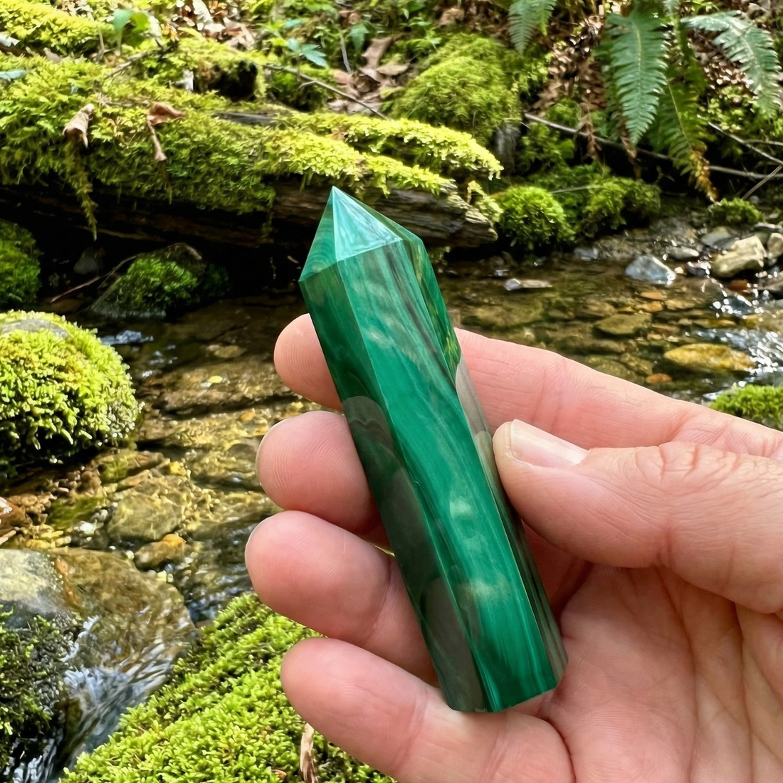 A banded green malachite point held in a hand beside a mossy forest stream