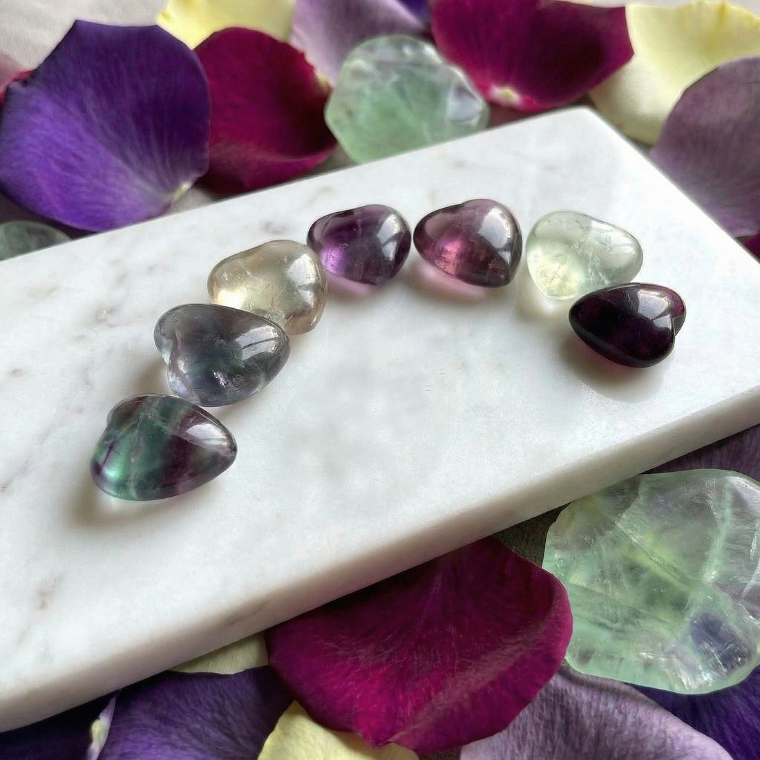 Seven polished fluorite hearts in a curved row on white marble, in green, purple, pale yellow and clear tones
