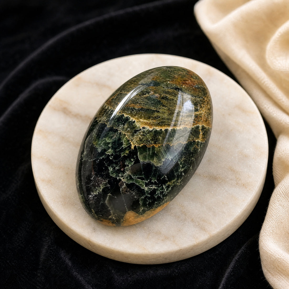 Polished ocean jasper palmstone on a cream marble dish, its glossy surface swirled with deep green, olive and golden ochre