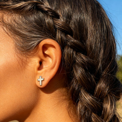 A small silver cross stud worn on the earlobe, beside a long dark braid in sunlight