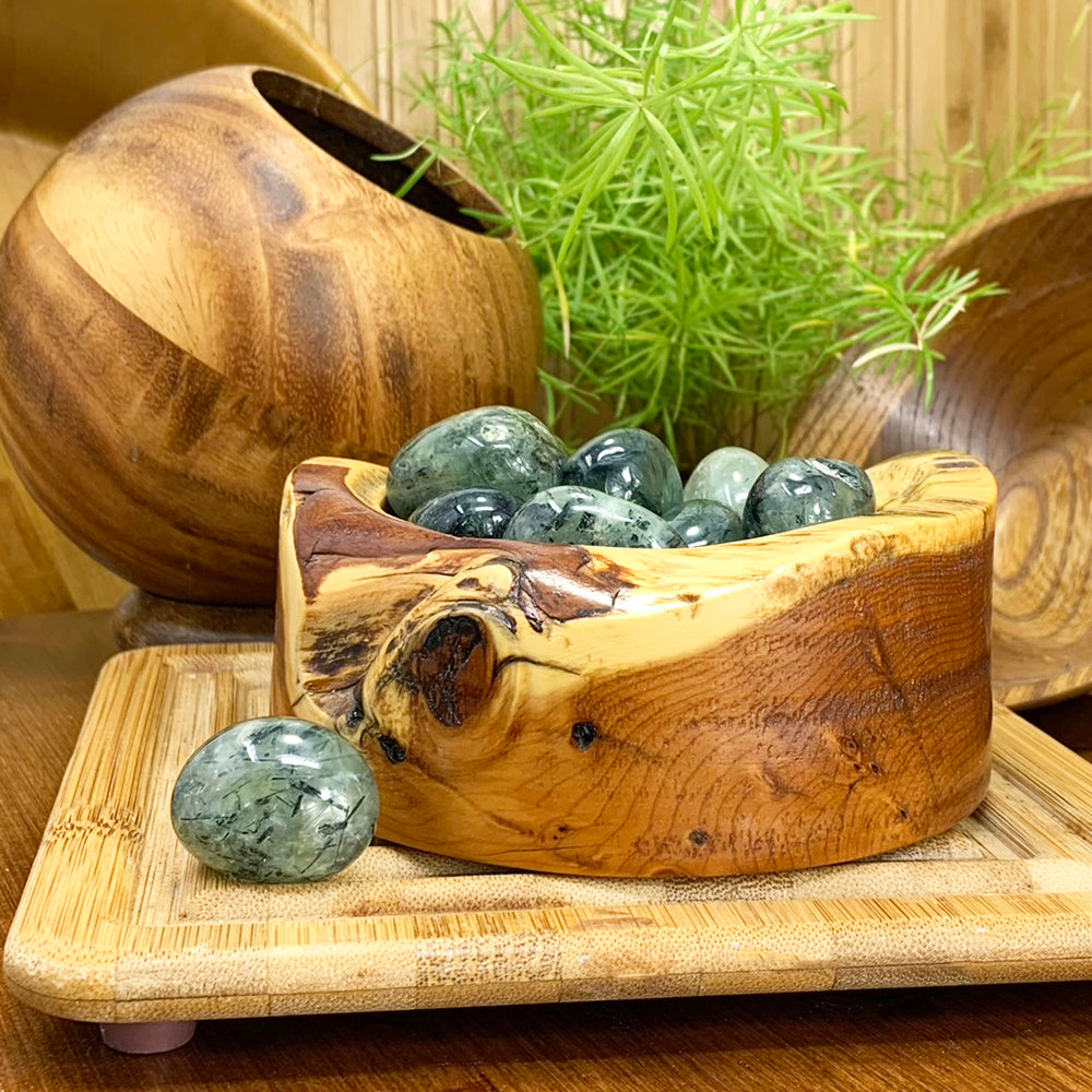 A tumbled prehnite ellipse in soft green with dark inclusions, resting in front of a wooden bowl of more prehnite stones
