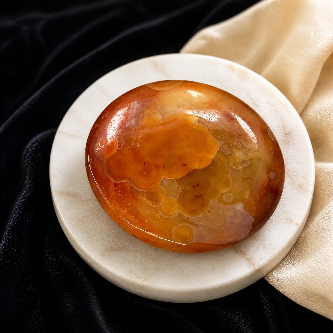 A polished oval carnelian palmstone in swirling orange, honey and red tones on a white marble dish