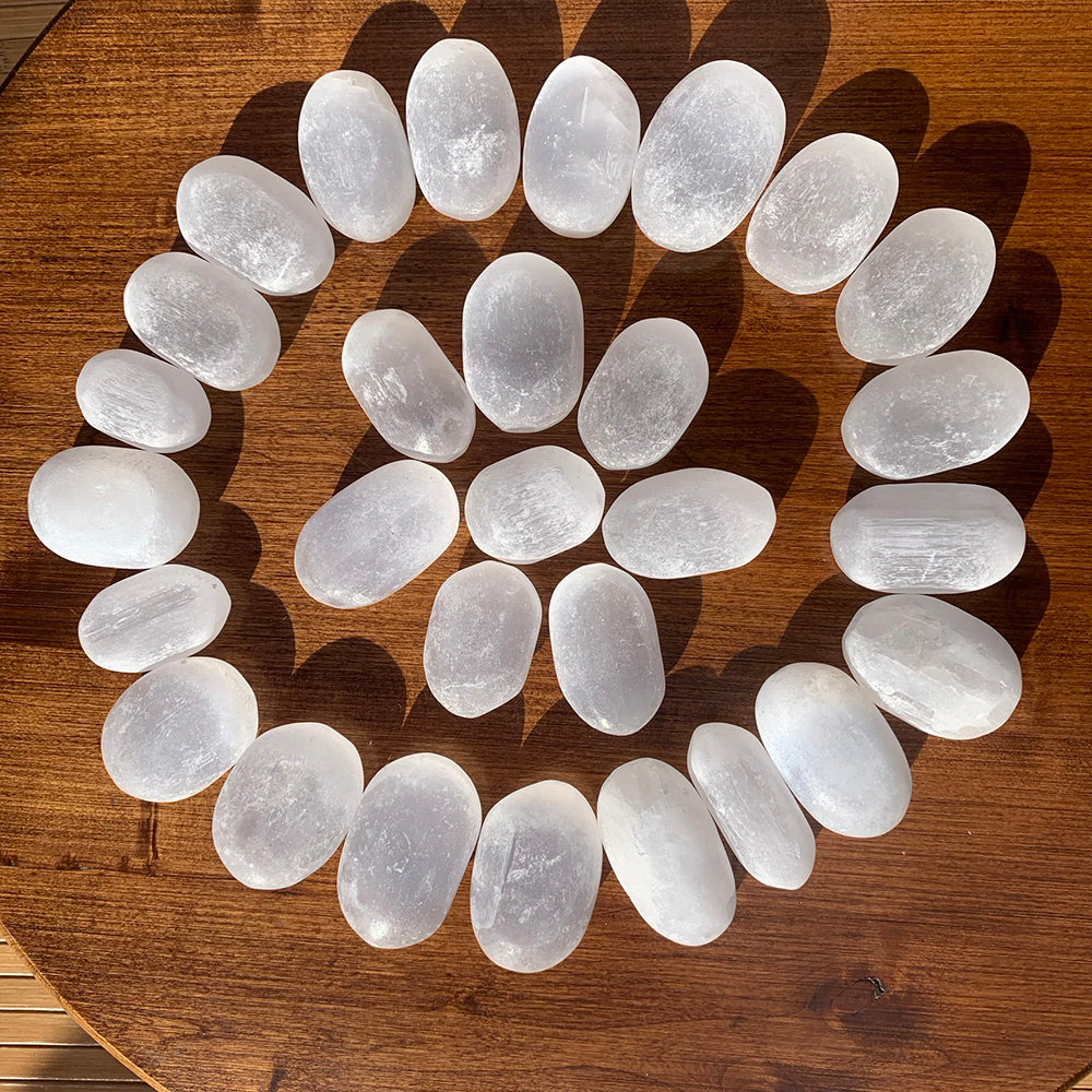 Dozens of selenite ellipse stones arranged in a spiral on a wooden table, each a slightly different shape with its own fibrous sheen