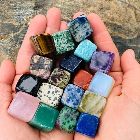 An open hand holding nineteen colourful tumbled crystal cubes, including a sodalite cube