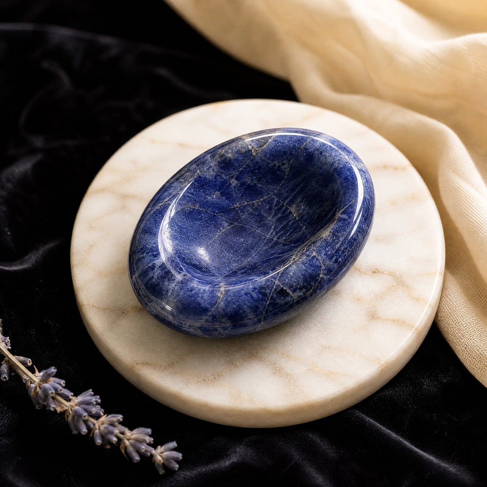 A polished royal blue sodalite worry stone with pale veining and a smooth thumb hollow, resting on a round marble dish