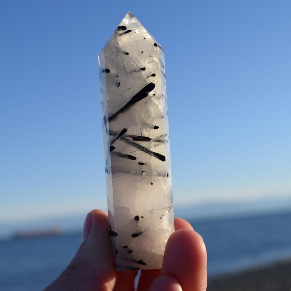 A tourmalinated quartz point with black tourmaline needles running through milky white quartz, held up against a blue sky